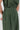 Close-up of a person wearing a dark olive green linen pleated jumpsuit or matching set, showing the waistband, side seam, and fabric texture against a light gray background.